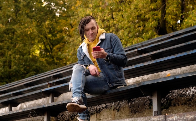 Jeune homme avec des dreadlocks naviguant sur un téléphone portable assis sur un vieux banc minable de gradin de rue