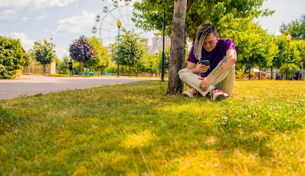 Un jeune homme avec des dreadlocks se repose à l'ombre d'un arbre et utilise un smartphone dans le parc Un beau mec à lunettes de soleil est assis sur l'herbe près d'un arbre par une journée ensoleillée