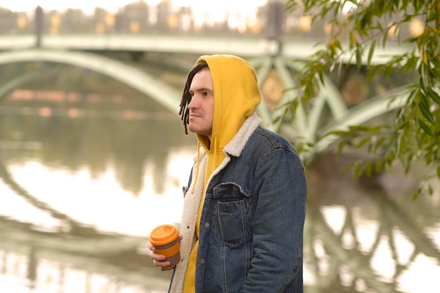 Jeune homme avec des dreadlocks tenant une tasse de café debout près du lac de la rivière sur fond de grand pont dans le parc de la ville