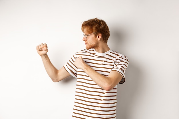 Jeune Homme Drôle Aux Cheveux Rouges Levant Les Poings Pour Le Combat, La Boxe De L'ombre Et L'air Sérieux Sur Le Côté Gauche, Debout Sur Fond Blanc