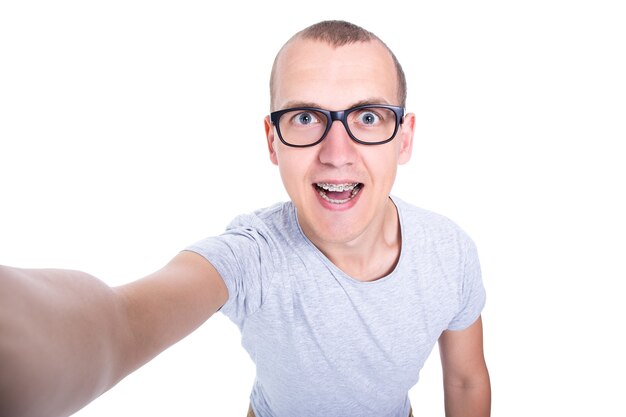 Jeune homme drôle dans des verres avec des accolades sur des dents prenant la photo de selfie d'isolement sur le fond blanc