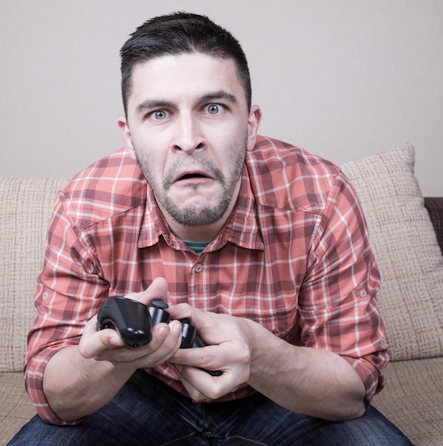 Jeune homme drôle jouant à des jeux vidéo assis sur un canapé et tenant une télécommande