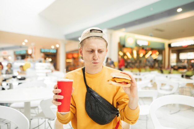 Un jeune homme drôle se tient avec un hamburger et un verre de cola à la main et a l'air contrarié par un fast-food