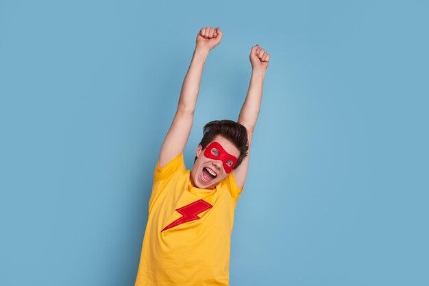 Jeune homme drôle en t-shirt de super-héros coloré et masque levant les bras et criant de stupéfaction sur fond bleu