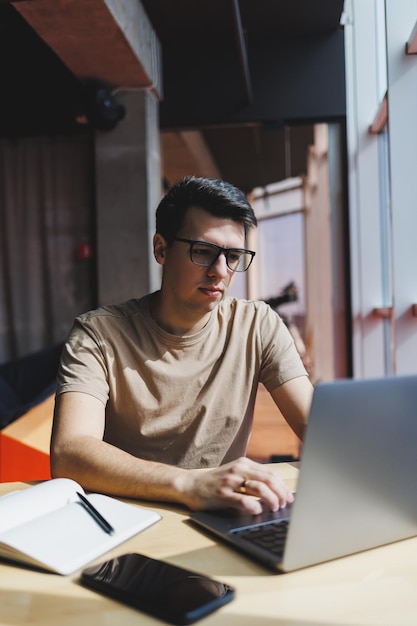 Jeune homme du millénaire assis à la table devant un ordinateur portable ouvert tout en étudiant en ligne Travail nomade numérique moderne en freelance sur netbook à l'intérieur d'un café