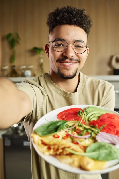 Photo un jeune homme du moyen-orient se photographie en tenant une assiette d'omelette et de légumes.