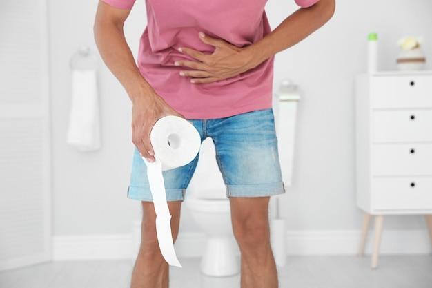 Photo jeune homme avec du papier hygiénique debout près de la cuvette des toilettes à la maison