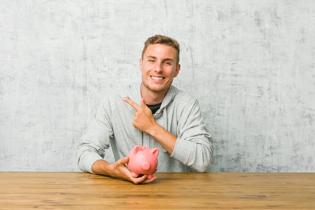 Jeune homme économiser de l'argent avec une tirelire souriant et pointant de côté, montrant quelque chose à l'espace vide.