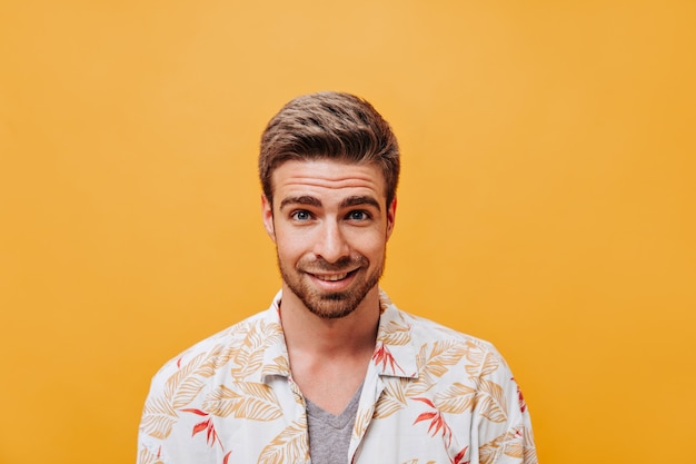 Jeune homme élégant à la barbe et aux yeux bleus en vêtements d'été imprimés blancs regardant la caméra sur un mur orange isolé