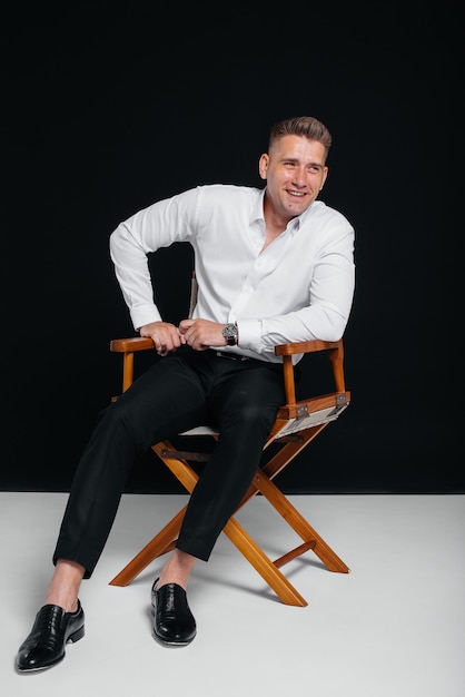 Un jeune homme élégant en chemise blanche est assis sur la chaise du réalisateur sur fond noir. Un bel homme d'affaires charismatique.