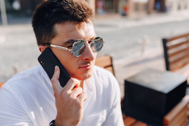 Jeune homme élégant en chemise avec téléphone sur banc par une chaude journée ensoleillée à l'extérieur