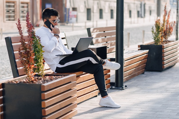 Jeune homme élégant en chemise avec téléphone et ordinateur portable sur un banc par une chaude journée ensoleillée à l'extérieur, freelance