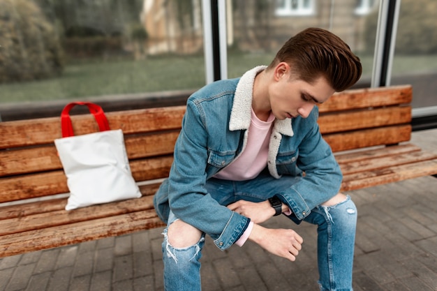 Un jeune homme élégant avec une coiffure dans des vêtements de jeans bleus décontractés vintage avec un sac en tissu à la mode est assis sur un banc en bois à un arrêt de bus en ville. Cool beau mec dans des vêtements de jeunesse en denim à la mode dans la rue.