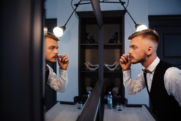 Jeune homme élégant corrigeant la moustache tout en regardant le miroir dans une pièce lumineuse Beau marié appréciant la préparation matinale pour la cérémonie de mariage