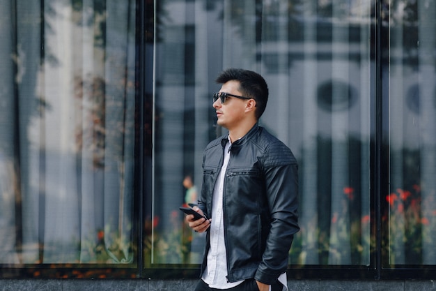 Jeune homme élégant dans des verres en veste de cuir noir avec téléphone sur fond de verre