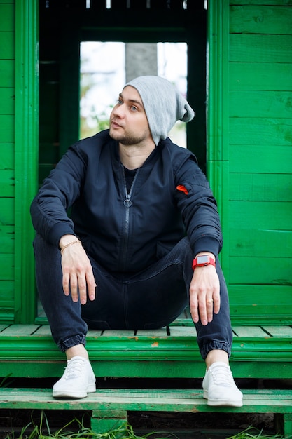 Jeune homme élégant dans une veste noire à la mode en jeans et chapeau posant par une journée ensoleillée près d'un bâtiment vert à l'extérieur. Mec moderne dans la rue. Style jeunesse