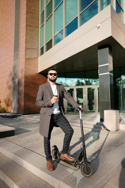 Jeune homme élégant dans des vêtements décontractés intelligents ayant un café à l'extérieur en se tenant debout sur un scooter électrique par l'architecture moderne