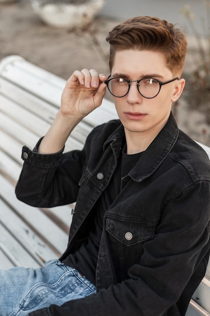 Jeune homme élégant dans des vêtements de jeans de mode redresse les lunettes vintage dans la rue. Cool modèle de gars européen dans des vêtements à la mode décontractés à la mode posant sur un banc blanc en bois dans la rue. Style décontracté