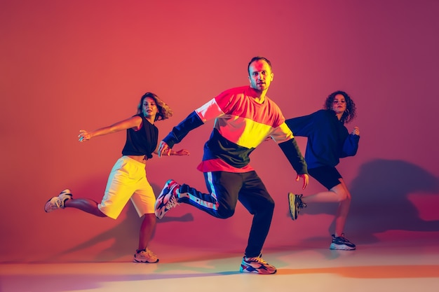 Jeune homme élégant et femmes dansant le hip-hop dans des vêtements lumineux sur fond dégradé à la salle de danse à la lumière du néon. Culture jeunesse, mouvement, style et mode, action, hip-hop. Portrait à la mode.