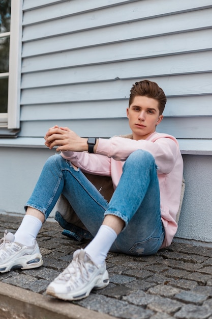 Jeune homme élégant en sweat rose à la mode en jean bleu tendance en baskets blanches est assis sur des carreaux de pierre près d'un bâtiment vintage dans la rue de la ville. Mec séduisant en vêtements de mode pour les jeunes à l'extérieur.