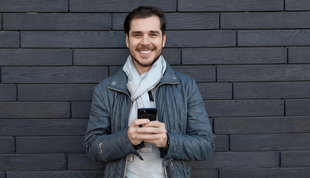 Un jeune homme élégant avec un téléphone dans ses mains regarde la caméra avec un large sourire sur fond noir