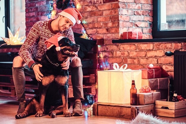 Un jeune homme élégant tenant une boîte-cadeau tout en étant assis avec son mignon chien dans un salon décoré au moment de Noël.