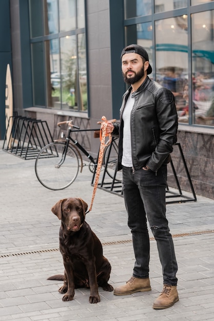 Jeune homme élégant en tenue décontractée debout sur trottoire pendant le refroidissement avec son animal mignon en milieu urbain