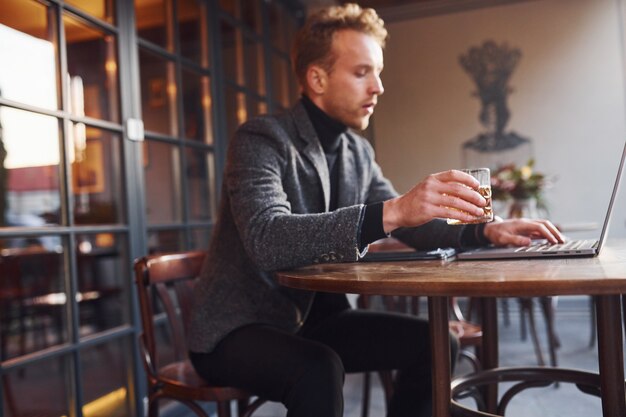 Un jeune homme élégant en tenue de soirée est assis dans un café avec son ordinateur portable et son verre d'alcool.