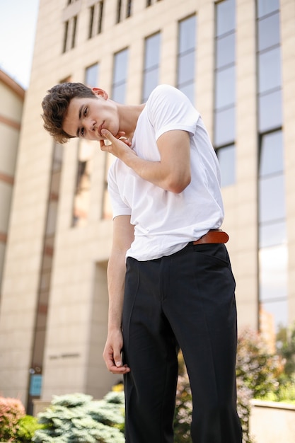 Un jeune homme élégant vêtu d'une chemise noire à la mode dans un t-shirt blanc élégant en pantalon noir avec une coiffure tendance repose près d'un centre d'affaires moderne. Mec séduisant dans la rue un jour d'été.