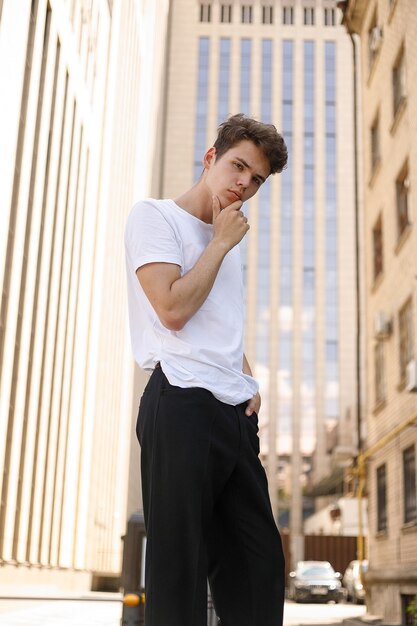 Un jeune homme élégant vêtu d'une chemise noire à la mode dans un t-shirt blanc élégant en pantalon noir avec une coiffure tendance repose près d'un centre d'affaires moderne. Mec séduisant dans la rue un jour d'été.