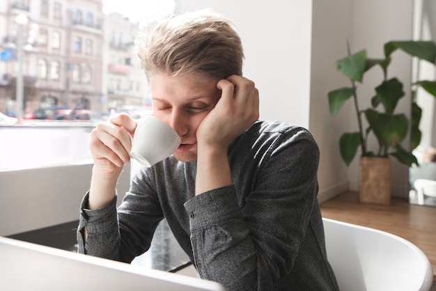 Un jeune homme endormi est assis le matin dans un café léger avec les yeux fermés dans un café et boit du café.