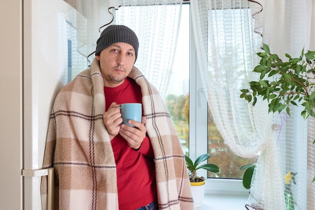 Un jeune homme enveloppé dans une couverture chaude se tient à la fenêtre de l'appartement avec une tasse de thé