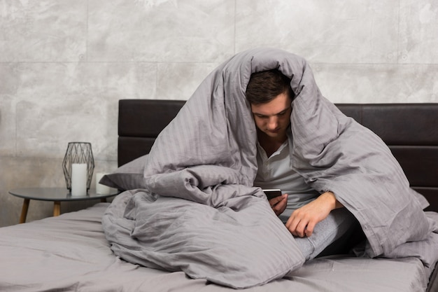 Jeune homme enveloppé dans une couverture et regardant son téléphone portable alors qu'il était assis dans un lit élégant aux couleurs grises et près d'une table de chevet avec des bougies dans une chambre de style loft