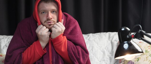 Photo un jeune homme enveloppé dans un plaid a l'air malade, malade, éternuant et toussant assis sur un canapé à la maison à l'intérieur. soins de santé et médecine, prévention des maladies, symptômes de maladies saisonnières et autoprotection. prospectus