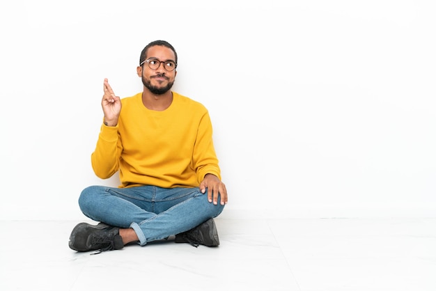 Jeune homme équatorien assis sur le sol isolé sur un mur blanc avec les doigts croisés et souhaitant le meilleur