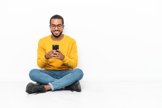 Jeune homme équatorien assis sur le sol isolé sur mur blanc envoyant un message avec le mobile