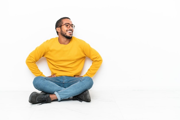 Jeune homme équatorien assis sur le sol isolé sur un mur blanc posant avec les bras à la hanche et souriant