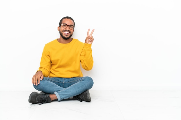 Jeune homme équatorien assis sur le sol isolé sur mur blanc souriant et montrant le signe de la victoire