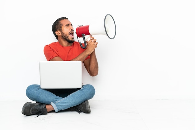 Jeune homme équatorien avec un ordinateur portable assis sur le sol isolé sur fond blanc criant à travers un mégaphone