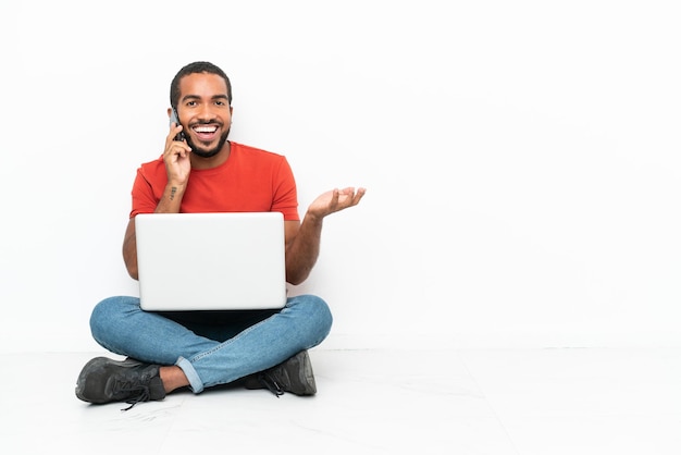 Jeune homme équatorien avec un ordinateur portable assis sur le sol isolé sur fond blanc en gardant une conversation avec le téléphone mobile avec quelqu'un