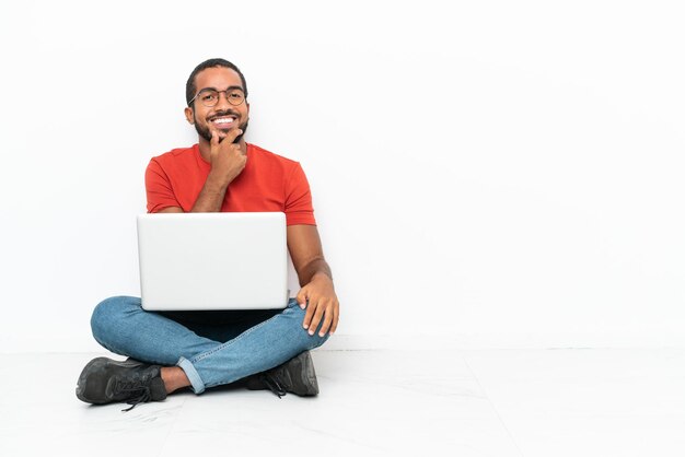 Jeune homme équatorien avec un ordinateur portable assis sur le sol isolé sur fond blanc avec des lunettes et souriant