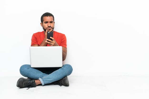 Jeune homme équatorien avec un ordinateur portable assis sur le sol isolé sur fond blanc pensant et envoyant un message