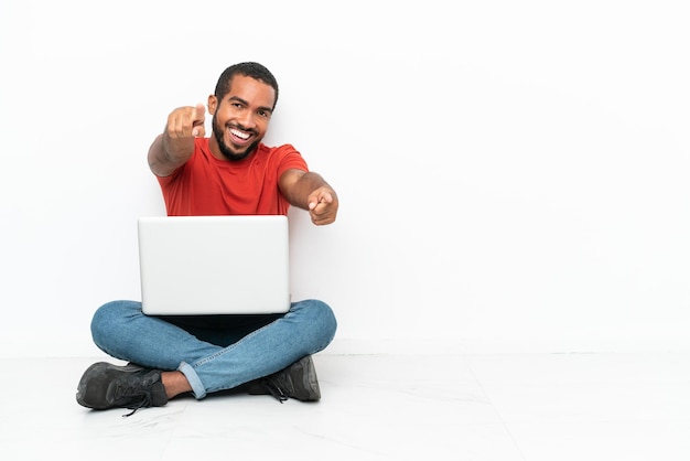 Jeune homme équatorien avec un ordinateur portable assis sur le sol isolé sur fond blanc pointe le doigt vers vous tout en souriant