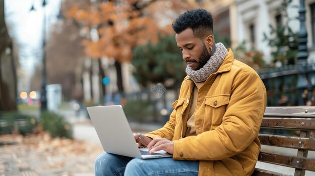 Un jeune homme est assis sur un banc dans le parc travaillant sur son ordinateur portable il porte une veste jaune jeans bleus et un foulard