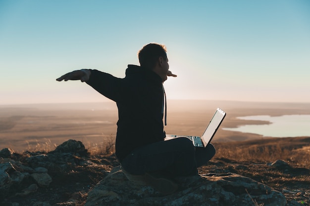 Un jeune homme est assis sur une colline et utilise un ordinateur portable. nomades numériques.