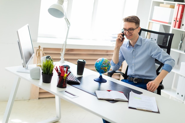 Un jeune homme est assis dans le bureau à un bureau d’ordinateur, parlant au téléphone et regardant le globe.