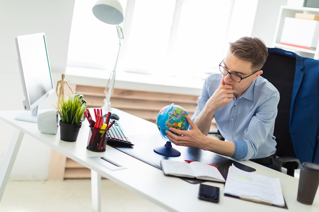 Un jeune homme est assis dans le bureau à un bureau d’ordinateur, regarde le globe et réfléchit.