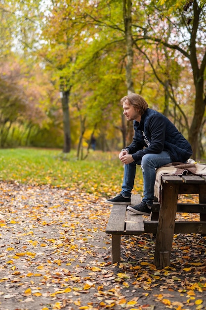 Le jeune homme est assis à l'extérieur en automne. Homme assis sur un banc dans un parc en automne. Heureux beau mec sur banc en bois