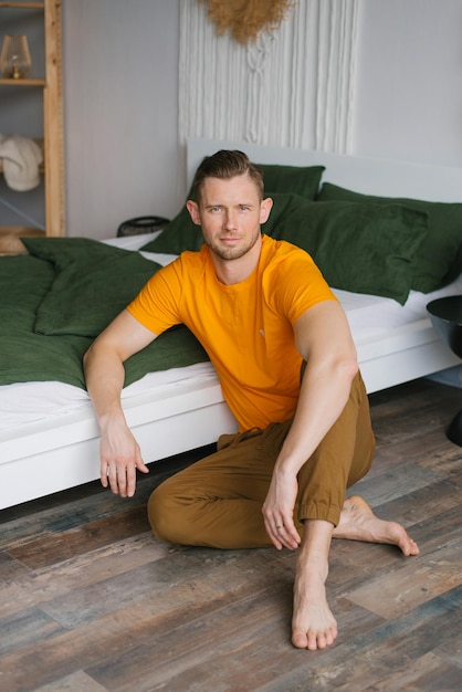 Jeune homme est assis à la maison sur le sol près du lit et regarde la caméra