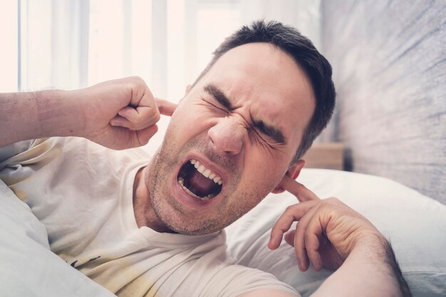 Le jeune homme est irrité par des bruits parasites pendant son sommeil Des voisins bruyants ou des réparations interfèrent avec le repos Le concept de bruit